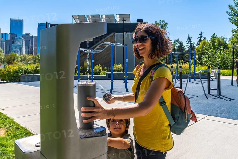 Tourist is filling reusable water bottle at a modern public drinking water fountain with calgary skyline in background, promoting sustainability and healthy lifestyle