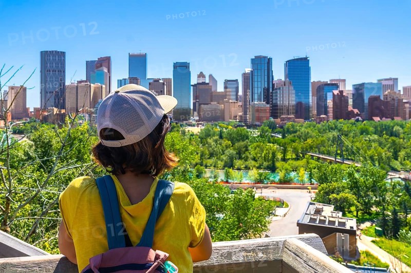 Tourist wearing a baseball cap and backpack enjoying a stunning view of the calgary skyline from a wooden viewpoint, surrounded by lush greenery on a sunny day