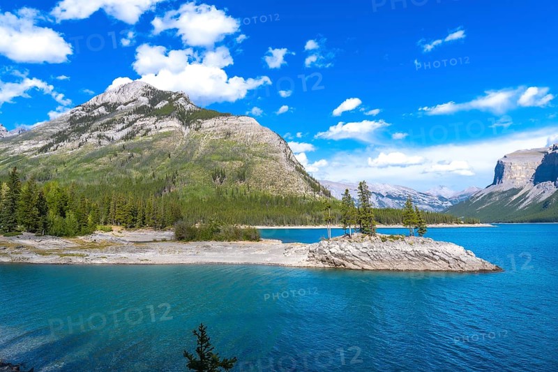 Turquoise water reflecting imposing mountains and a rocky islet with coniferous trees under a bright blue sky with clouds in lake minnewanka, banff national park, canadian rockies, alberta, canada