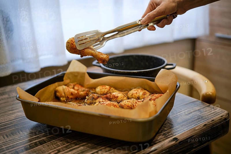 A hand uses tongs to pick up a roasted chicken drumstick from a tray in a kitchen. The tray sits on a wooden table with light coming through a window in the background