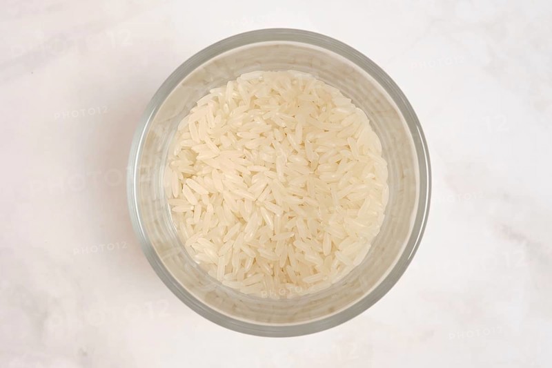 A clear bowl contains uncooked white rice. The rice is fluffy and dry, waiting to be cooked. The bowl is positioned on a light-colored surface, showing a simple kitchen setup