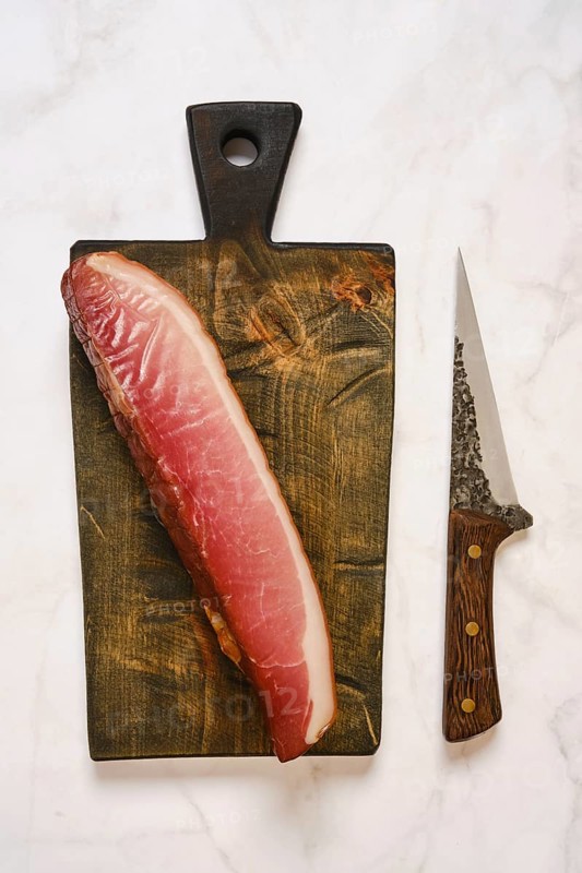Cured parma ham on a wooden cutting board. A sharp knife is placed next to it. This scene is set in a kitchen, ideal for meal preparation or cooking activities