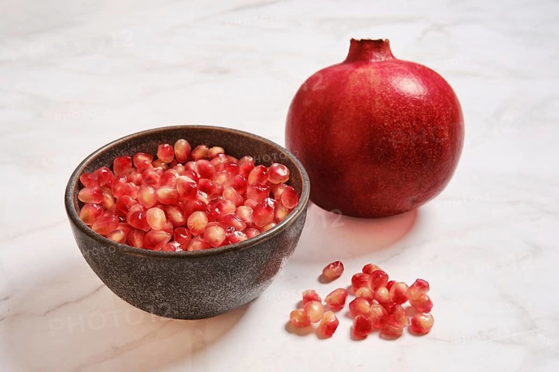 A bowl contains bright pomegranate seeds next to a whole pomegranate. The bowl is placed on a light countertop with a few scattered seeds around. The scene shows fresh fruit