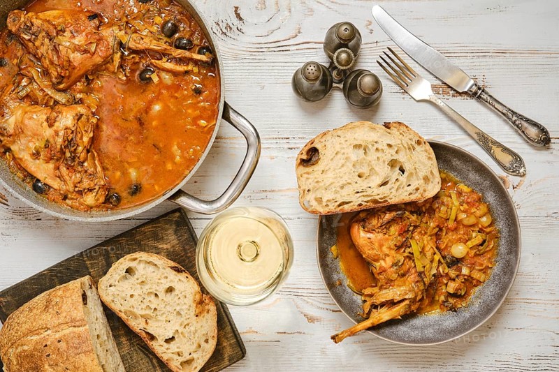 A rabbit stew with olives and vegetables sits in a pan next to a bowl. A piece of bread lies beside the bowl. A glass of white wine stands nearby. The setting shows a simple dining scene