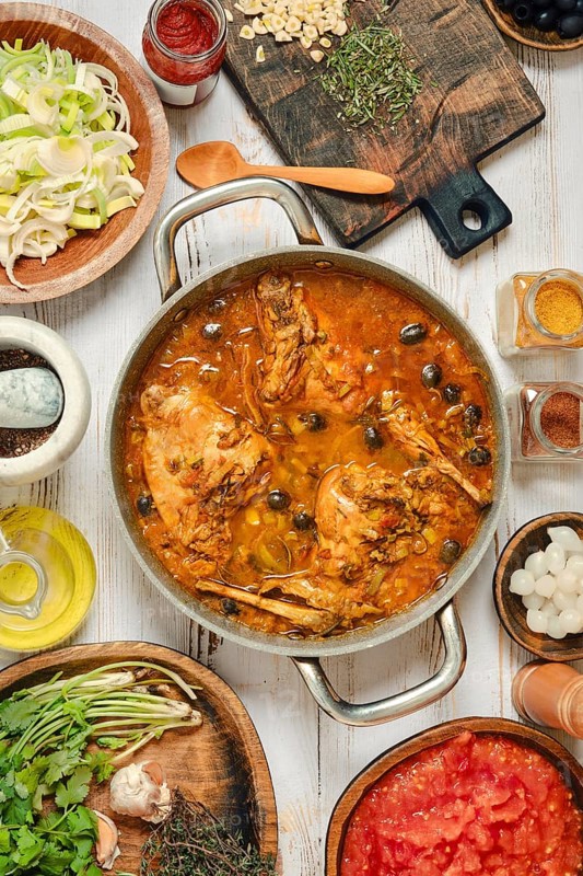 A large pot holds a simmering stew made with rabbit meat, vegetables, and a variety of spices. Surrounding the pot are bowls of ingredients like herbs, chopped tomatoes, and onions ready for use