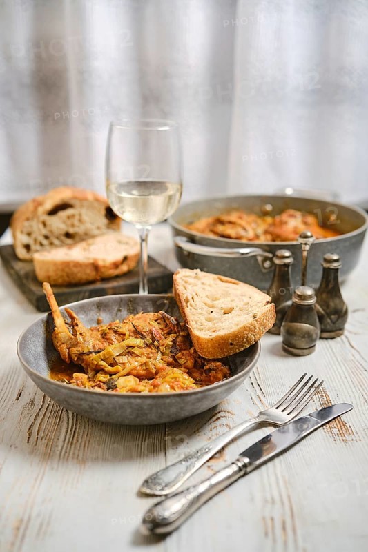 A dinner table is prepared with a bowl of food, a glass of white wine, and slices of bread. Other servings are placed nearby, creating a warm atmosphere for a meal