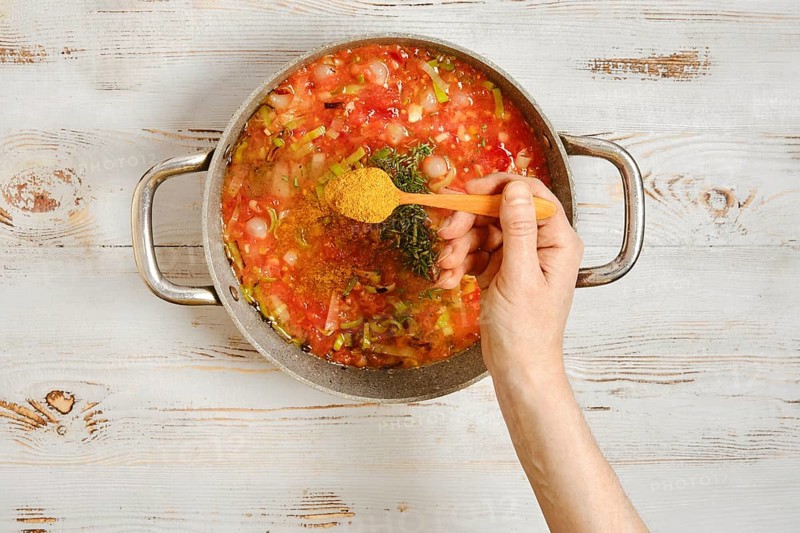 A hand sprinkles spice in a pot with chopped vegetables and a rich tomato base. Spices are added to enhance the flavor. The kitchen has a light wooden background