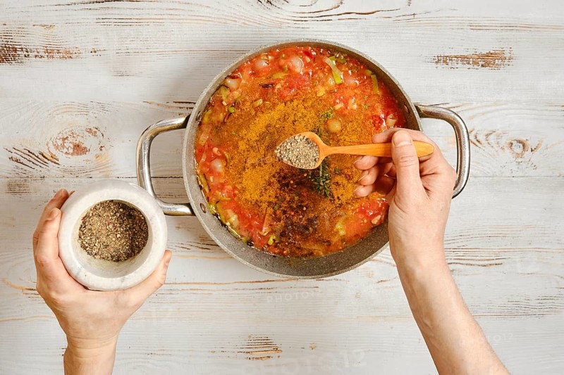 A person adds pepper to a pot of vegetables and tomatoes. The hands hold a wooden spoon and a mortar. Fresh ingredients are mixed as the meal is being prepared in a cozy kitchen