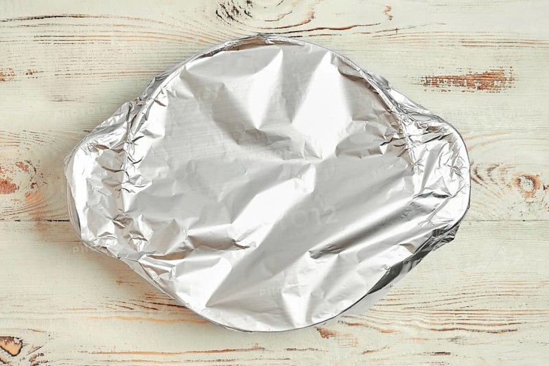A roasting pot is covered with aluminum foil placed on a wooden kitchen table. The foil prevents excess browning while baking. It is part of a cooking activity in a home setting