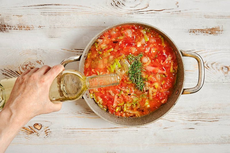 A person pours white wine into a pot filled with chopped tomatoes and vegetables. The pot sits on a wooden table. Fresh herbs are added to enhance the flavor