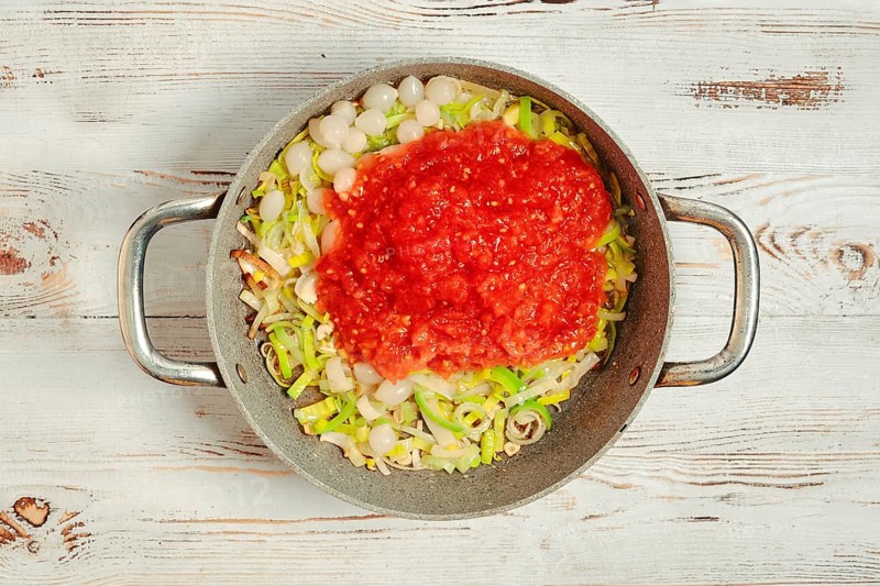On a wooden surface, a pan holds chopped leek and pearl onions. A layer of crushed tomato sits on top of the vegetables, ready for cooking