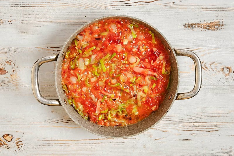 A pot filled with a mixture of chopped vegetables and tomatoes on a wooden kitchen counter. The food is being prepared for a meal, showcasing various colors and textures