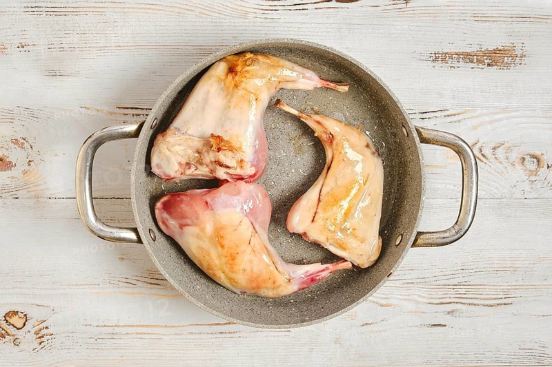 Three raw rabbit legs are browning in a metal pot on a wooden surface. The scene shows the preparation of meat for cooking. The pot has a simple design with a shiny finish