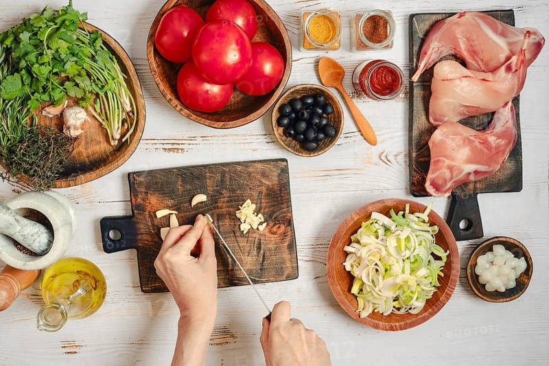 A person is chopping garlic while other ingredients are ready for cooking. Various spices sit nearby for seasoning