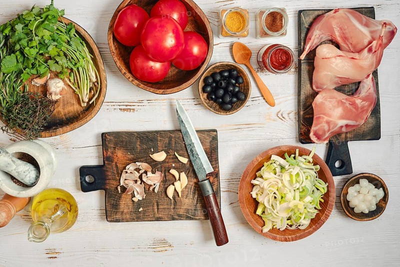 Fresh tomatoes, herbs, and spices are laid out on wooden boards in a kitchen. A knife and cutting board sit nearby, along with garlic, rabbit, and other food items ready for cooking