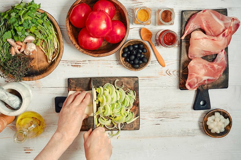 Fresh vegetables, meats, and spices are arranged on a table. Someone is chopping onions with a knife while tomatoes and other ingredients are nearby. This scene shows meal preparation in a kitchen