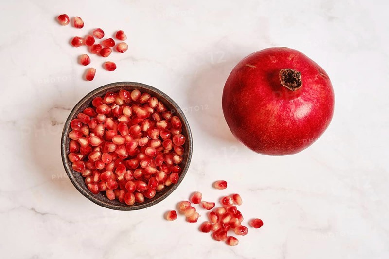 A round red pomegranate sits next to a bowl filled with bright red seeds on a white marble surface. The natural shine of the pomegranate stands out against the simple background