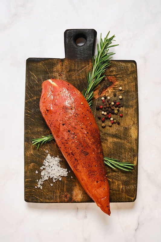Cured turkey meat on a wooden cutting board. Around it are herbs, spices, and salt. The setting is a kitchen countertop. The colors of the ingredients blend well together. Careful preparation is visible