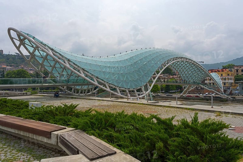 Wavy, modern glass bridge across parkland with hills in the background, Peace Bridge, Rike Park, Tbilisi, Kura River, Georgia