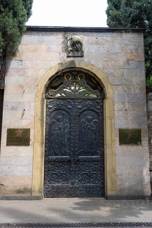 Decorated iron gate in a stone wall with plaques and surrounded by trees, entrance to the Georgian Patriarchate, the headquarters of the Georgian Orthodox Church, Tbilisi, Georgia