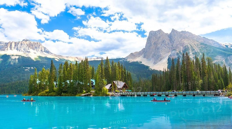 Canoeing tourists glide across the turquoise waters of emerald lake, framed by a wooden bridge, lush forest, and snow capped mountains in banff national park