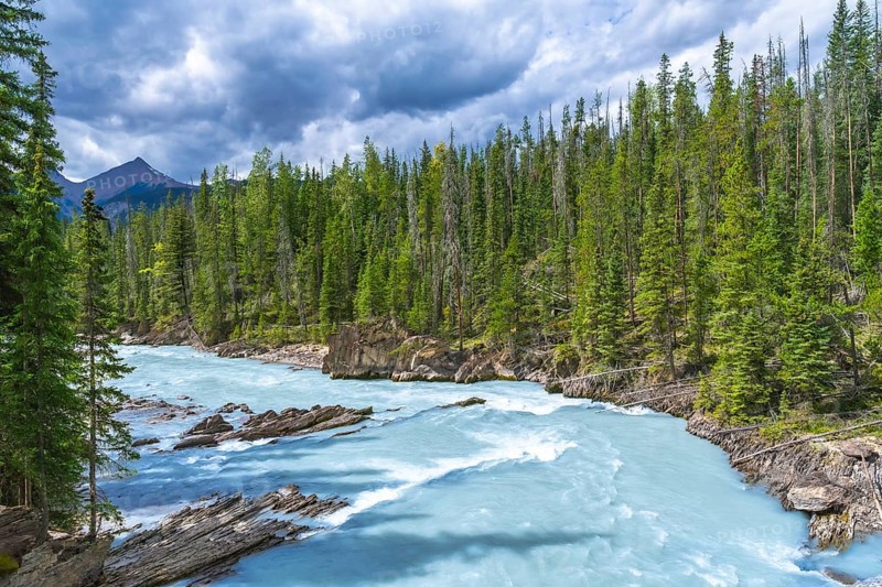 Turquoise water flows through the bow river natural bridge in banff national park, alberta, canada, surrounded by lush green pine trees and a cloudy sky