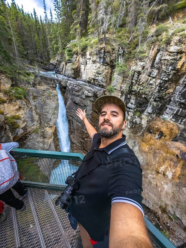 Tourist taking a selfie with outstretched arm at johnston canyon lower falls, showcasing the stunning waterfall, canyon, and lush forest in banff national park, alberta, canada, during a sunny day