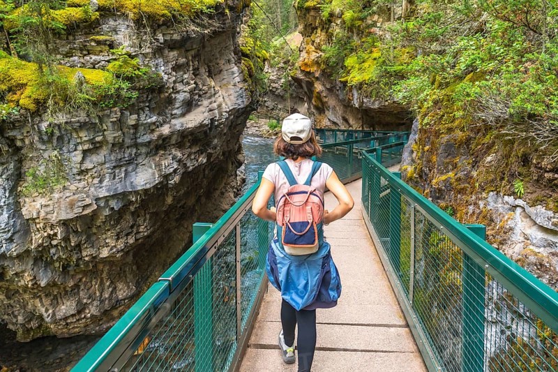 Female tourist with backpack walking on metal catwalk enjoying the view of johnston canyon and the bow river in banff national park, canadian rockies, alberta, canada, during summer
