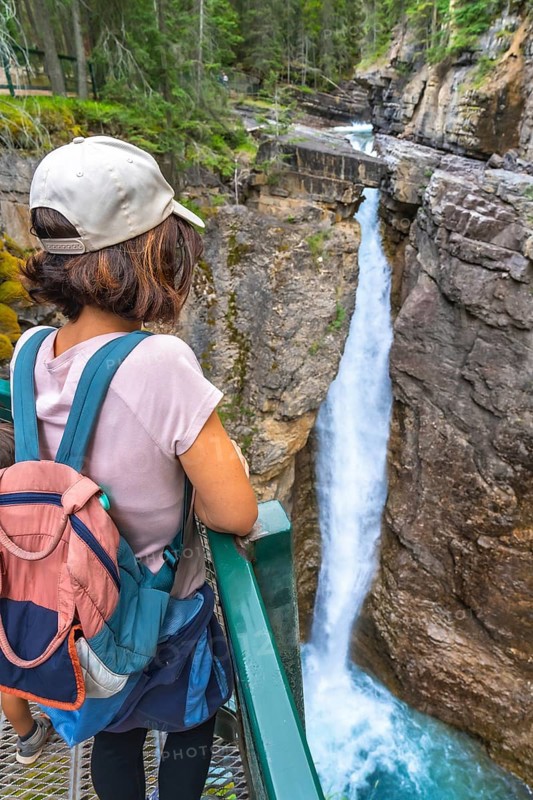 Female tourist carrying a backpack and baby carrier, enjoying the stunning view of a waterfall cascading into the turquoise waters of johnston canyon in banff national park
