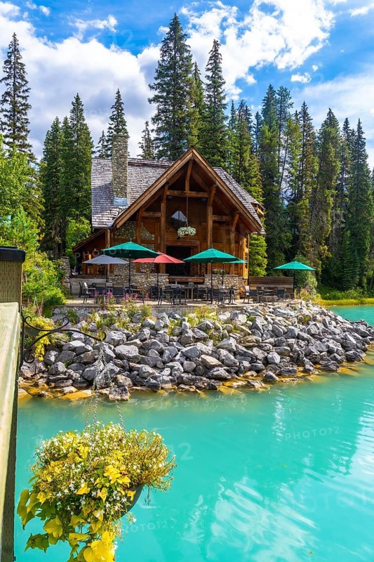 Wooden lodge with patio furniture and umbrellas reflecting in turquoise water on the shore of emerald lake with hanging flower basket in the foreground, yoho national park, british columbia, canada