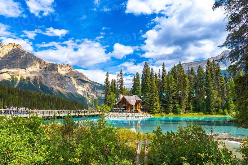 Wooden lodge reflecting on turquoise water with tourists walking on a bridge, surrounded by mountains and forest on a sunny day at emerald lake in yoho national park, canada