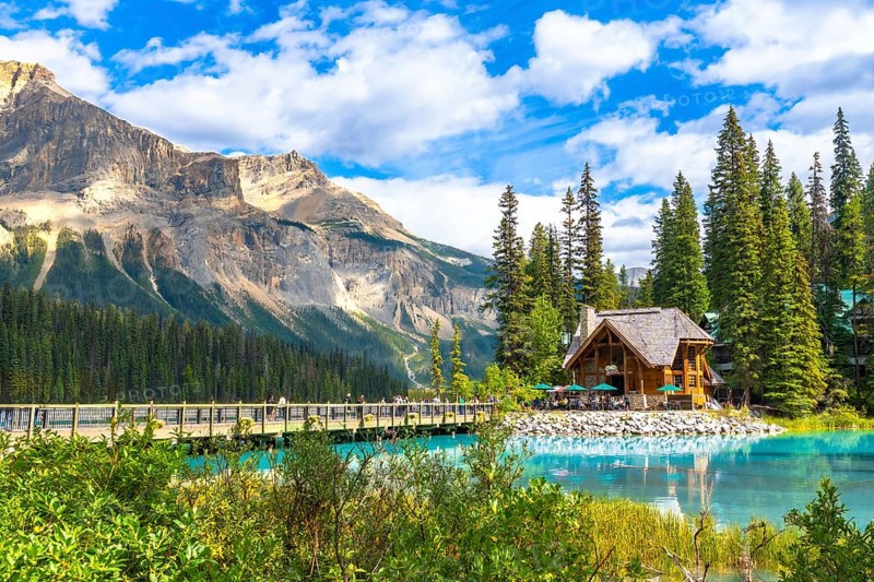 Wooden lodge sitting on the shore of a turquoise lake reflecting the surrounding lush vegetation and mountain range under a blue sky with clouds