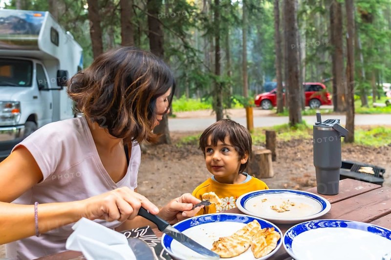 Mother giving grilled food to her child while having lunch at a campsite in the canadian rockies, banff national park, alberta, with their campervan parked in the background