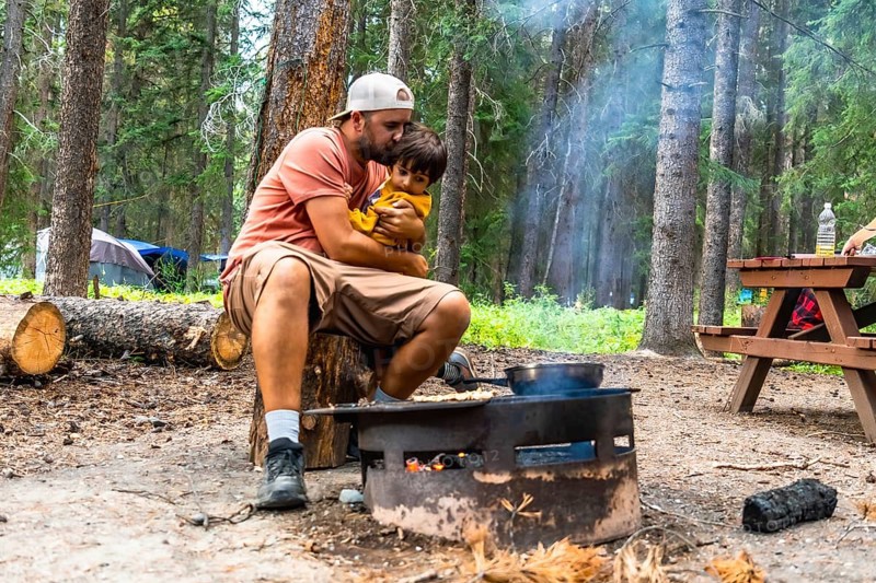 Father showing affection to his son while cooking breakfast on campfire during a camping trip in banff national park, canadian rockies, alberta
