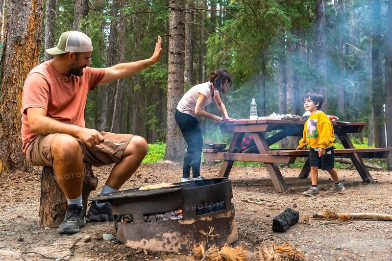 Family camping and grilling while bonding in the breathtaking scenery of banff national park, with a father waving, a mother preparing food, and a child walking near the campfire