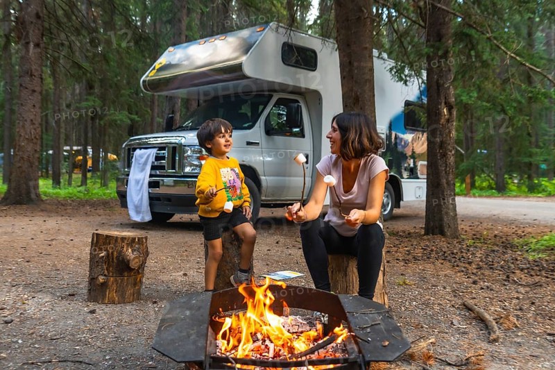 Mother and son roasting marshmallows over a campfire, enjoying quality time together during a camping trip in banff national park, with their campervan parked nearby in the serene forest