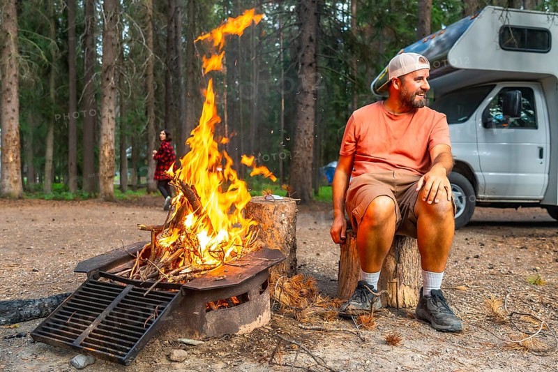 Camper enjoying warmth of campfire in banff national park, canadian rockies, with campervan parked nearby and another camper walking in the background