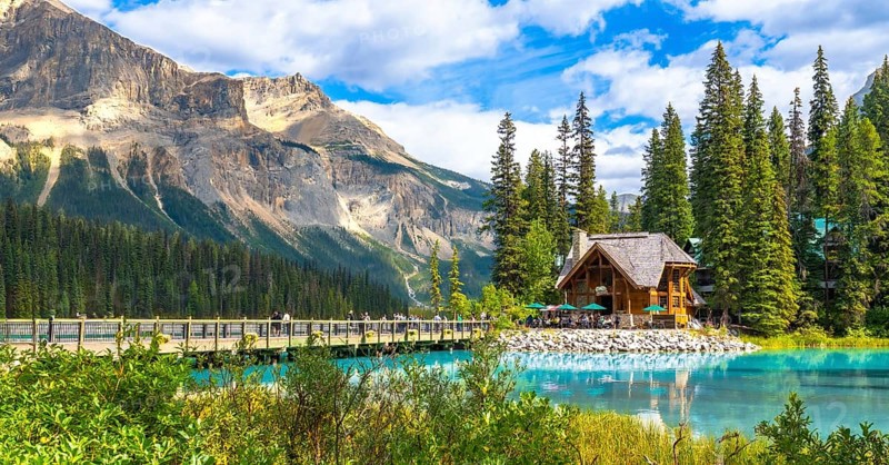 Wooden lodge sitting on the shore of a turquoise lake reflecting in the calm water with tourists walking on a bridge and a mountain in the background in yoho national park