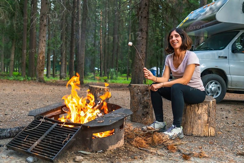 Female tourist roasting marshmallows over a campfire in a campground in banff national park, next to her camper van, enjoying the peace and quiet of nature