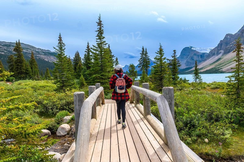 Female tourist with backpack walking on a wooden bridge at bow lake surrounded by lush vegetation and mountains. In banff national park. Canadian rockies. Alberta. Canada. During a cloudy summer day