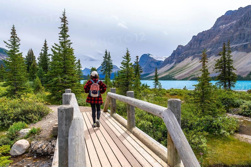 Female tourist walking on a wooden bridge and enjoying the breathtaking view of bow lake and surrounding mountains in banff national park, alberta, canada, during a cloudy summer day