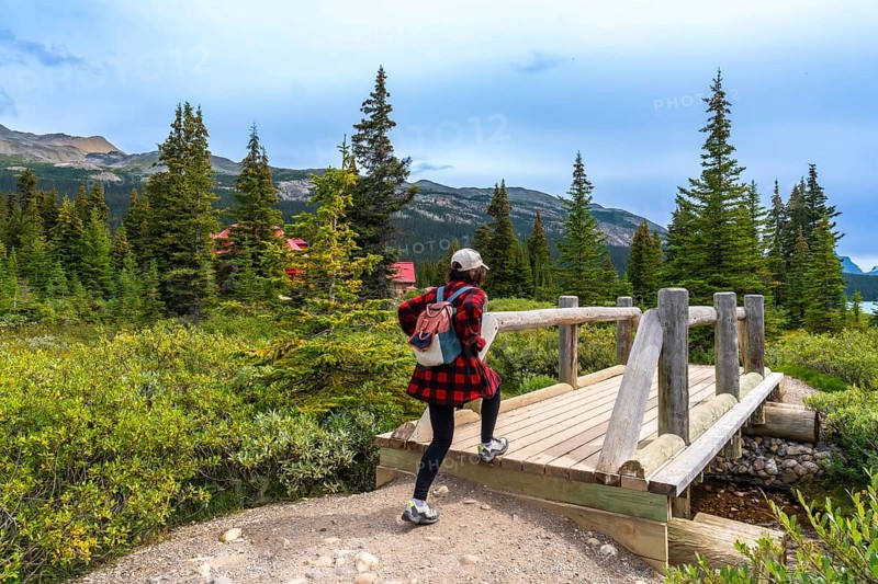Female tourist walking across a wooden bridge at bow lake, framed by a red roofed building, coniferous trees, and majestic mountains under a cloudy sky in banff national park