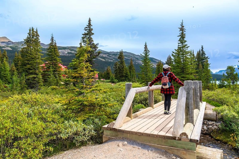 Tourist standing on a wooden bridge, admiring the breathtaking view of bow lake and surrounding mountains in banff national park, alberta, canada, with a red lodge among the trees