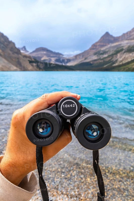 Hand holding binoculars, focusing on turquoise waters of bow lake surrounded by majestic mountains and glaciers in banff national park, alberta, canada