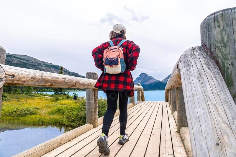 Female tourist walking on wooden bridge enjoying the scenic view of bow lake and surrounding mountains in banff national park, canadian rockies, alberta, canada, during a cloudy day