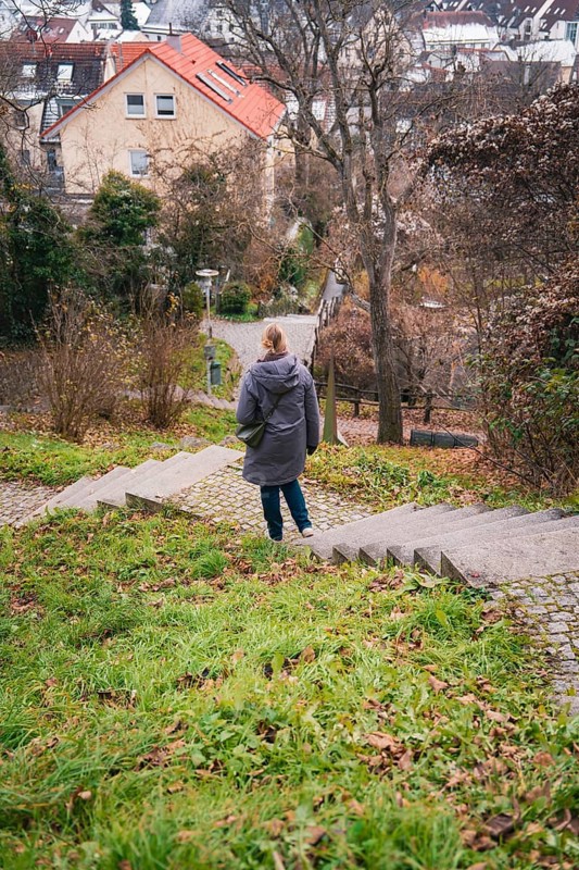 A woman in a cloak walks down an overgrown staircase surrounded by trees and houses, Herrenberg, Böblingen district, Germany