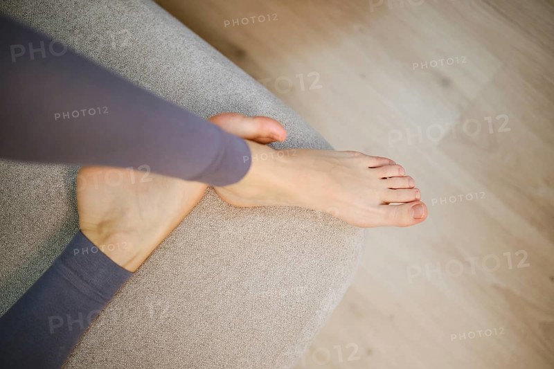 Barefoot female legs resting on a sofa in a home setting during a daytime. The focus is on the foot and leg area