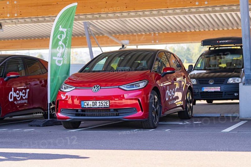 Red car sharing car under covered parking lot next to green banner and modern cityscape, ID3, Deer e-Carsharing, Germany