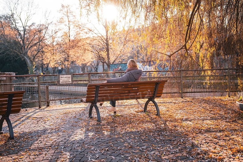 Woman sitting on a park bench in autumn surrounded by golden foliage at sunset, Stadtgarten Pforzheim, Germany