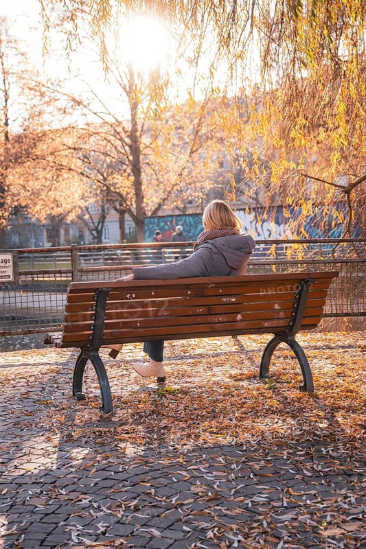 Woman sitting on park bench surrounded by autumn trees in sunlight, Stadtgarten Pforzheim, Germany